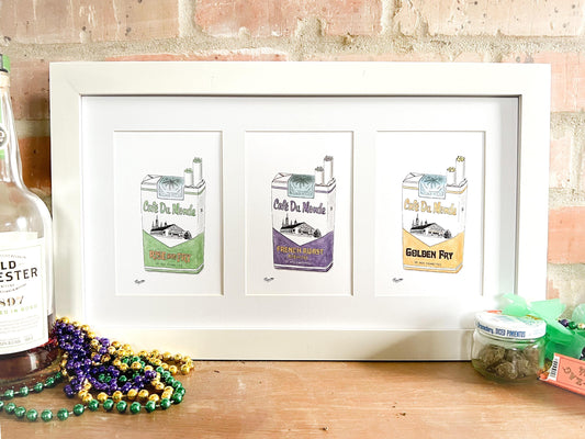 A product photo of the Cafe Du Monde Collection set of art prints. A single horizontal frame with 3 separate 5x7 mat openings displays all three versions of the Cafe DuMondeJazz Cigarettes collection. As order in frame: the green "Rise and Fry", the purple "French Roast", and the gold "Golden Fry". The frame rests against a brick wall on a mantle with Mardi Gras colored beads and a bottle of bourbon on the left while a small repurposed pimento jar holding a green herb and rolling papers sit on the left.