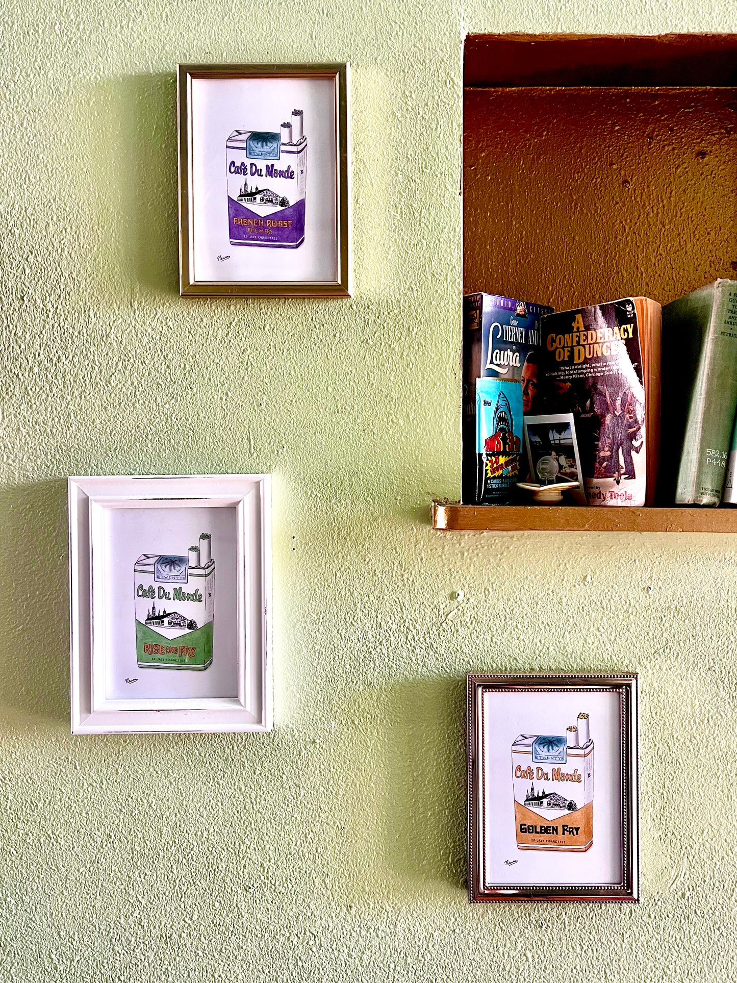 The collection of all three 5x7 Cafe Du Monde Jazz Cigarette packs are individually framed and hung on a wall next to a book shelf displaying the novel "A Confederacy of Dunces" by John Kennedy Toole.