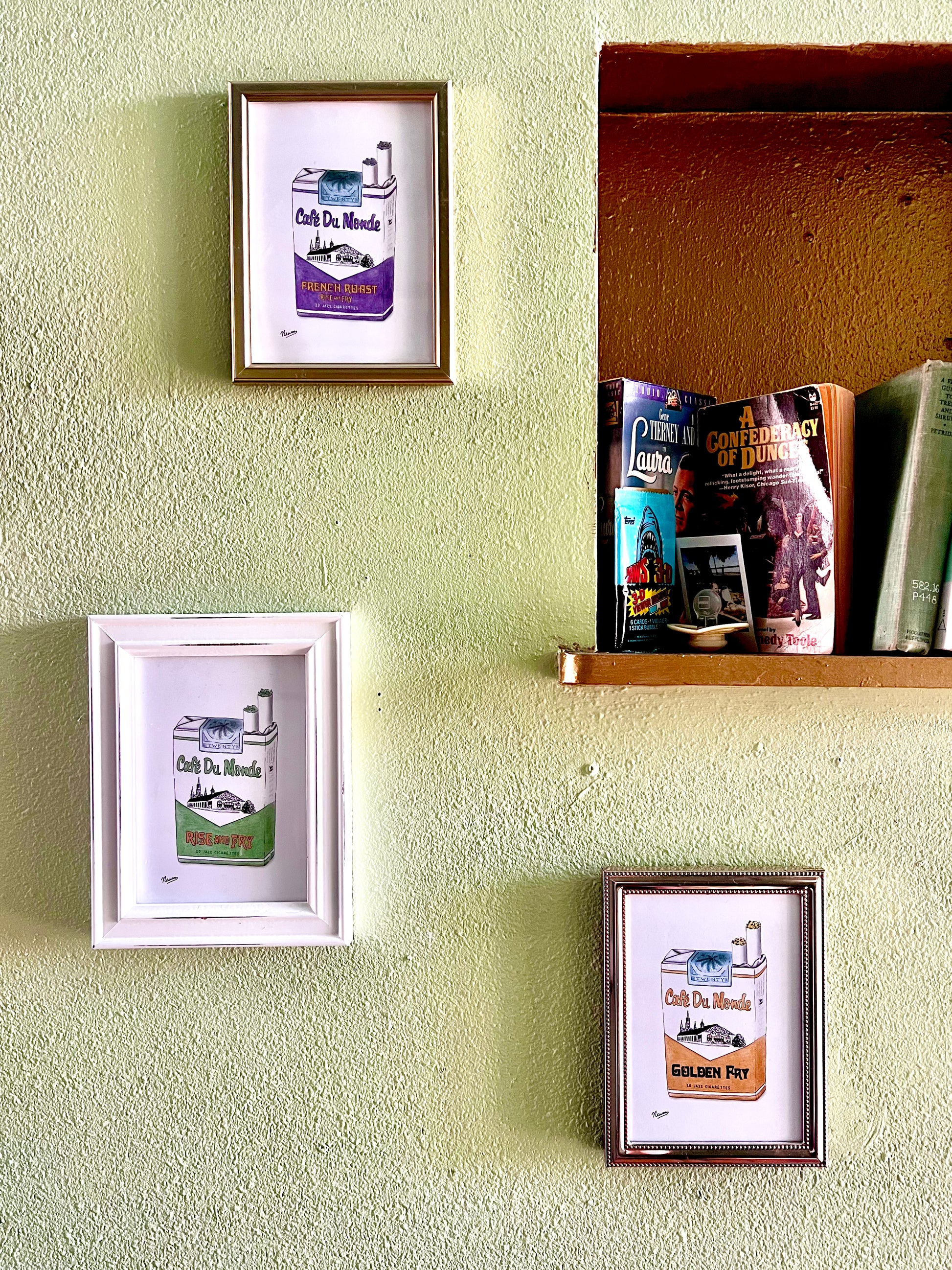 The collection of all three 5x7 Cafe Du Monde Jazz Cigarette packs are individually framed and hung on a wall next to a book shelf displaying the novel "A Confederacy of Dunces" by John Kennedy Toole.