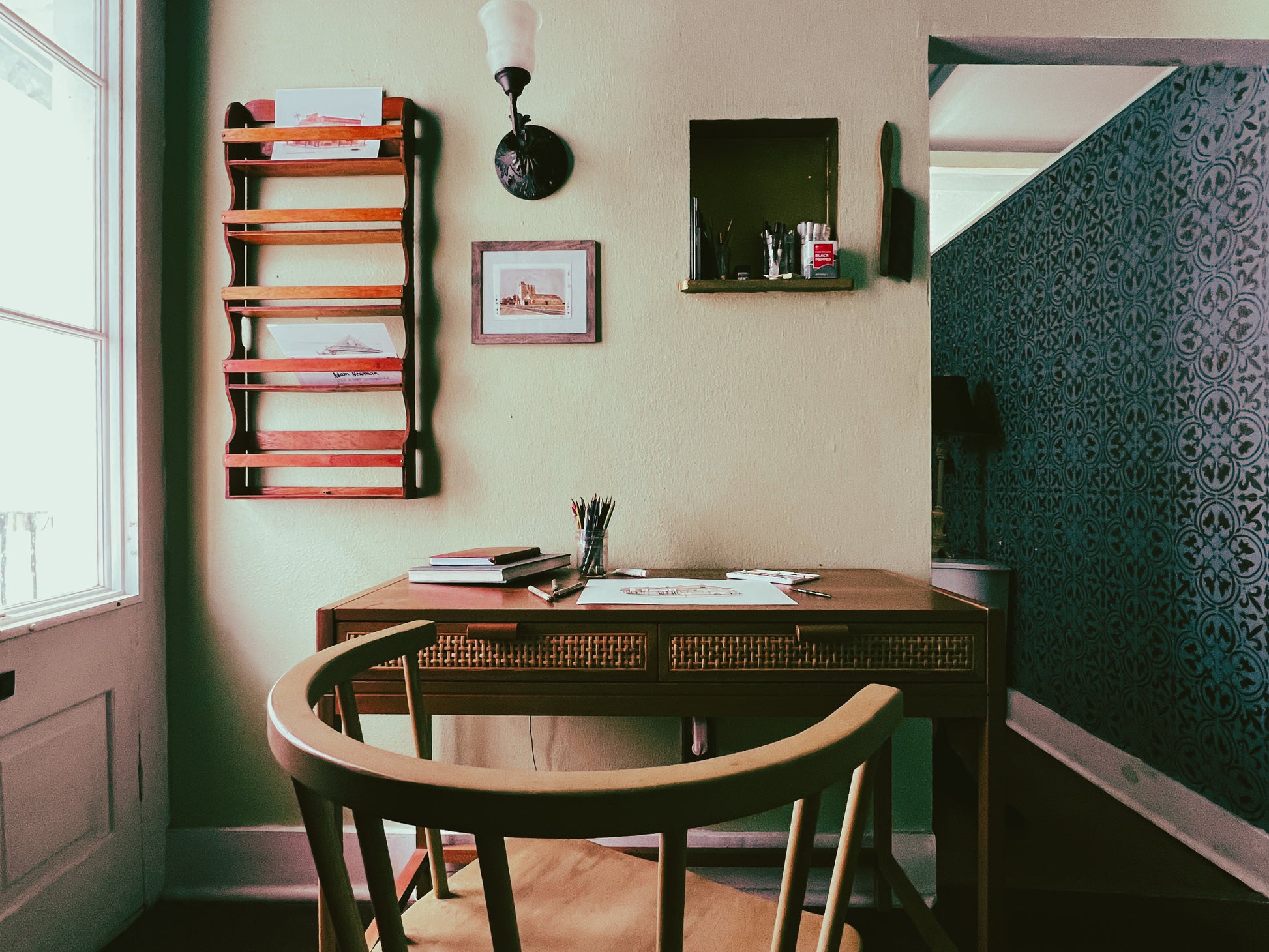 A view of the artist's studio. An enclave in the wall holds pencils and brushes with a soft brush for wiping dust hangs just to the right, and on the left a sconce, small framed painting by the artist, and a mail rack. A door leads to a balcony overlooking the French Quarter on the left lets in an abundance of natural light. Finally the focal point is the artist's desk & chair pulled away having a finished piece lying flat on the surface along with pencils, brushes, pens, paint, and several sketchbooks.
