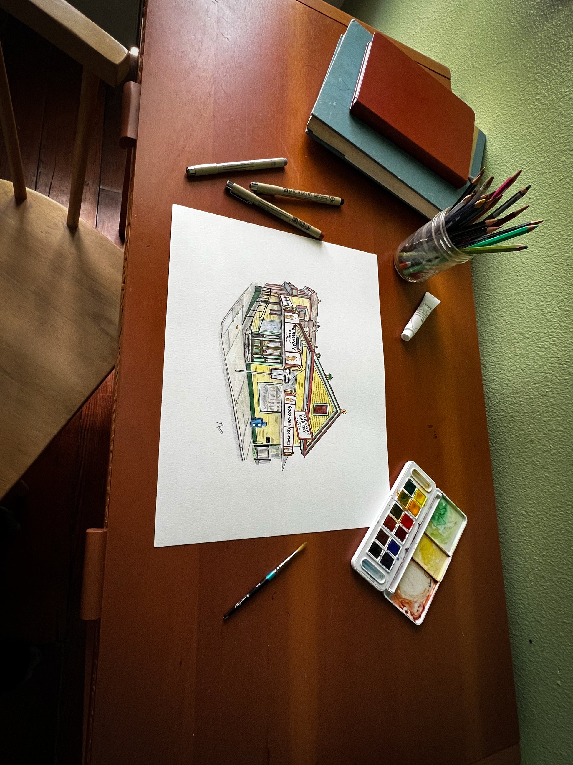 The finished Parkway Bakery piece on paper rests on a desk with several sketchbooks, pens, a tray of watercolor paint, a brush, a tube of gouache, and a small jar full of color pencils.