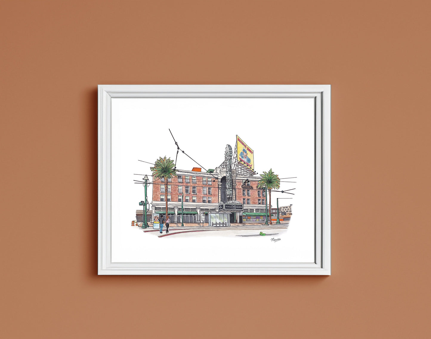 A detail of the Saenger Theater art print in a frame. The finished piece sits in a white frame on a warm brown wall. The frame matches the white background on which the theater floats in the artwork.