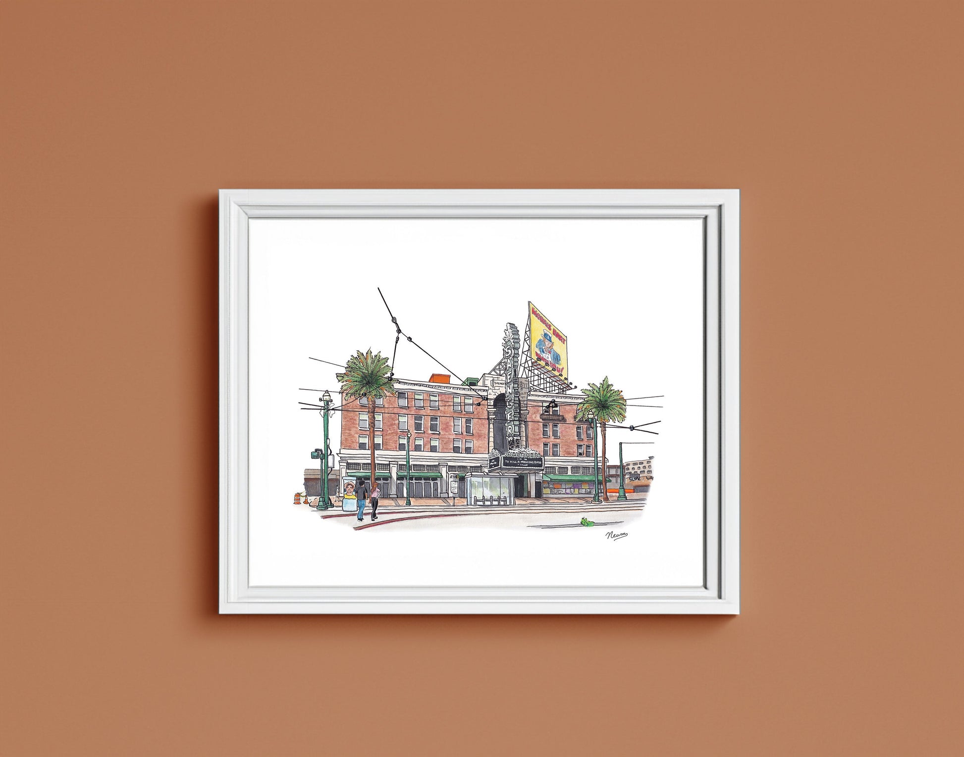 A detail of the Saenger Theater art print in a frame. The finished piece sits in a white frame on a warm brown wall. The frame matches the white background on which the theater floats in the artwork.