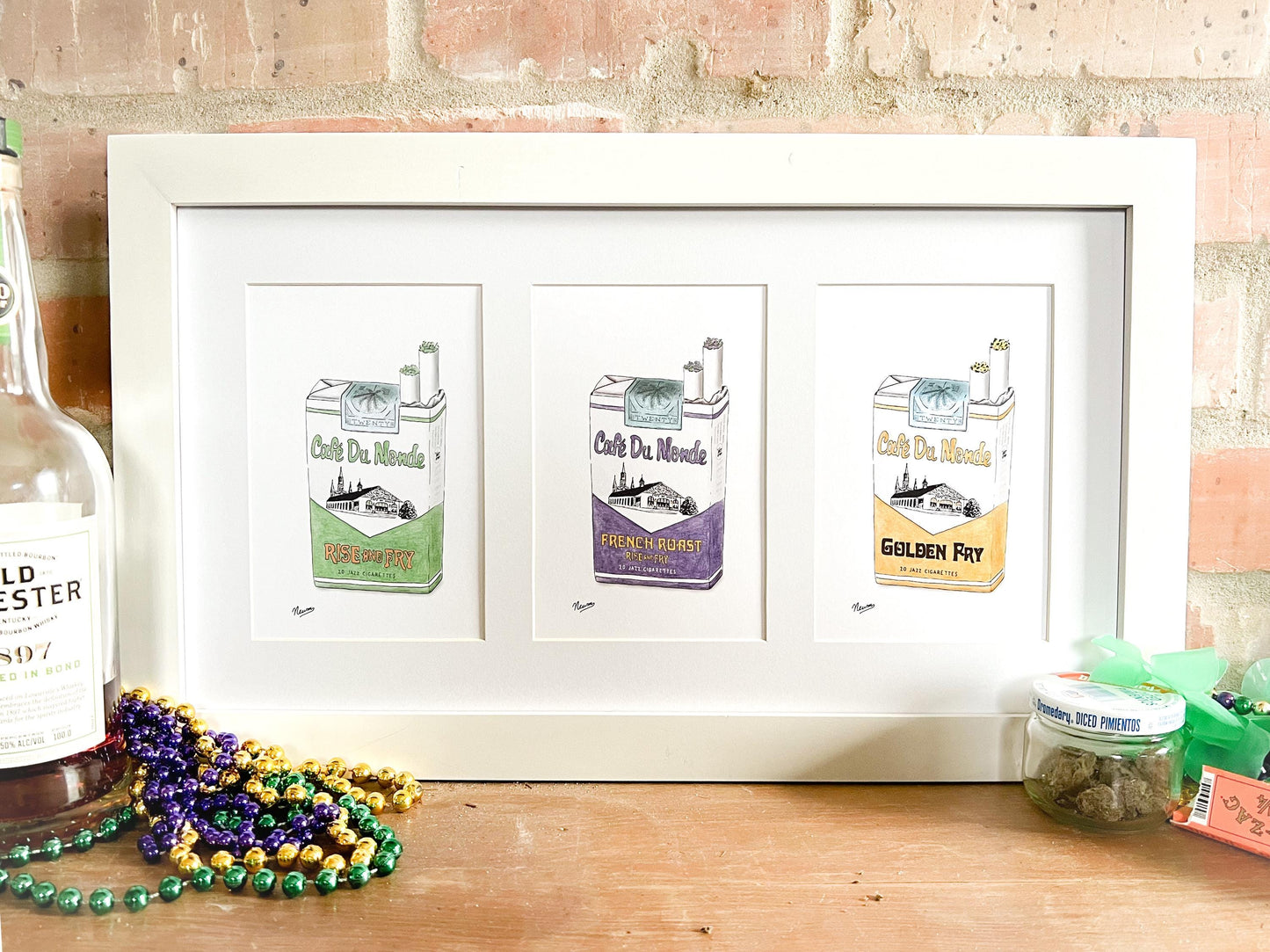 A product photo of the Cafe Du Monde Collection set of art prints. A single horizontal frame with 3 separate 5x7 mat openings displays all three versions of the Cafe DuMondeJazz Cigarettes collection. As order in frame: the green "Rise and Fry", the purple "French Roast", and the gold "Golden Fry". The frame rests against a brick wall on a mantle with Mardi Gras colored beads and a bottle of bourbon on the left while a small repurposed pimento jar holding a green herb and rolling papers sit on the left.